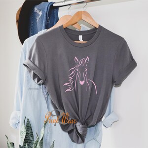 K&ouml;nnte beinhalten: Ein graues T-Shirt mit einem rosafarbenen Pferdesilhouetten-Design. Das Shirt h&auml;ngt an einem Holzb&uuml;gel.