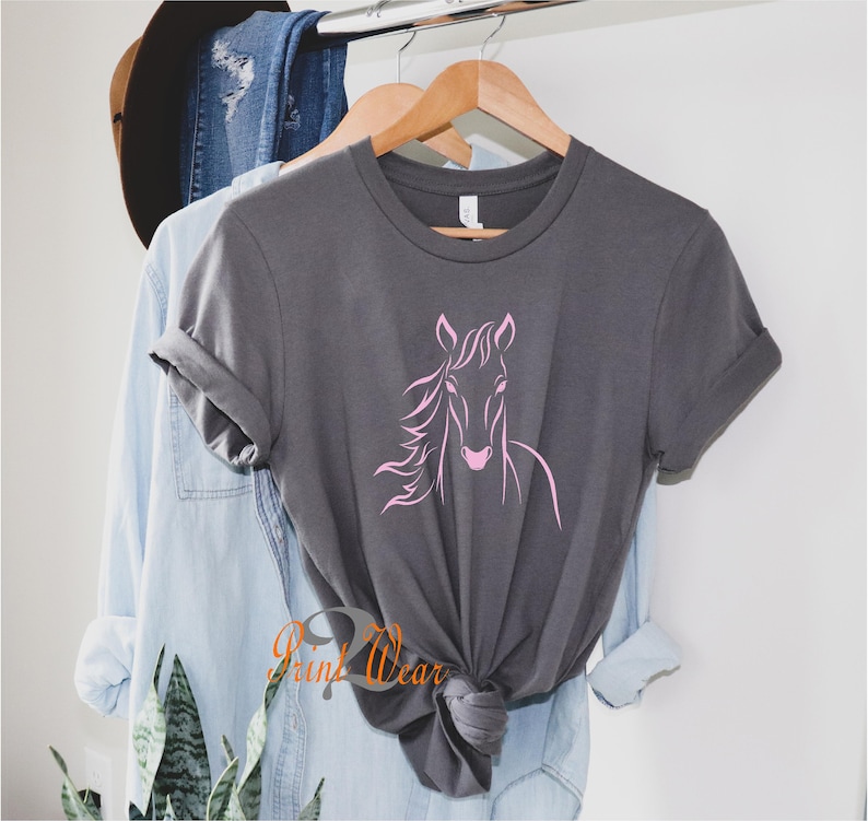 K&ouml;nnte beinhalten: Ein graues T-Shirt mit einem rosafarbenen Pferdesilhouetten-Design. Das Shirt h&auml;ngt an einem Holzb&uuml;gel.