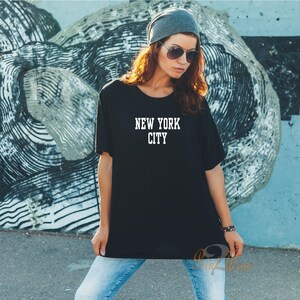 Puede incluir: Una camiseta negra con el texto "NEW YORK CITY" impreso en blanco en la parte delantera. La camiseta la lleva puesta una persona con el pelo largo y casta&ntilde;o y una boina gris.