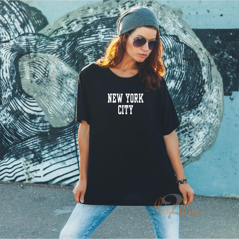 Puede incluir: Una camiseta negra con el texto "NEW YORK CITY" impreso en blanco en la parte delantera. La camiseta la lleva puesta una persona con el pelo largo y casta&ntilde;o y una boina gris.