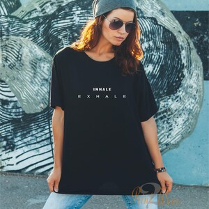 Może przedstawiać: Czarny t-shirt z kr&oacute;tkim rękawem z białym napisem INHALE EXHALE na piersi. Koszulka ma okrągły dekolt i luźny kr&oacute;j, zestawiona z jasnoniebieskimi dżinsami, szarą czapką typu beanie oraz okularami przeciwsłonecznymi z ciemnymi szkłami.