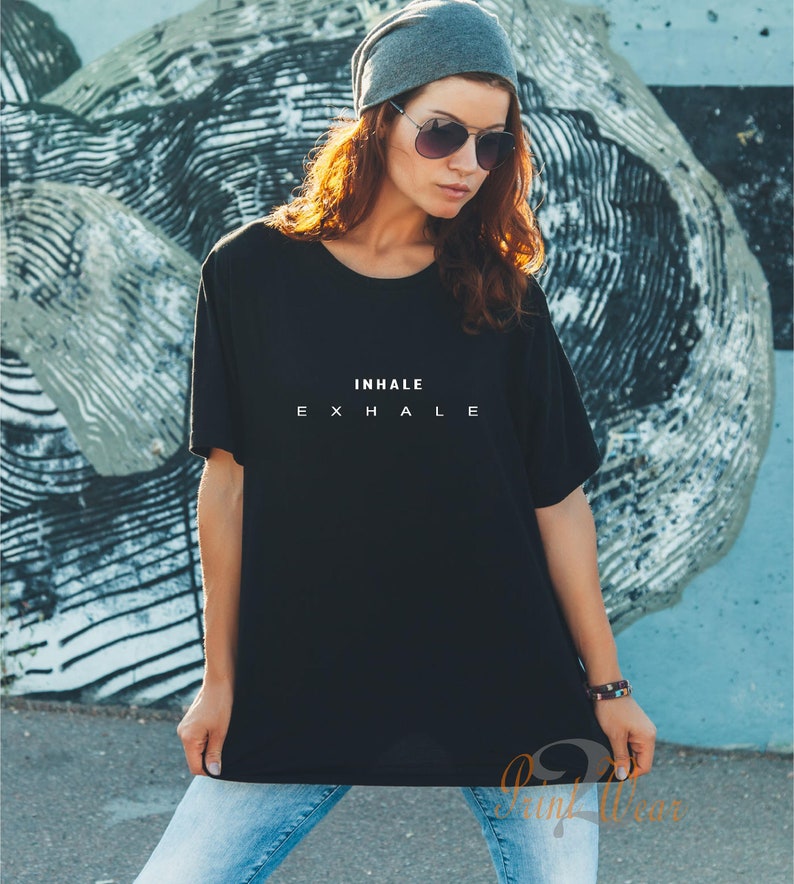 Może przedstawiać: Czarny t-shirt z kr&oacute;tkim rękawem z białym napisem INHALE EXHALE na piersi. Koszulka ma okrągły dekolt i luźny kr&oacute;j, zestawiona z jasnoniebieskimi dżinsami, szarą czapką typu beanie oraz okularami przeciwsłonecznymi z ciemnymi szkłami.
