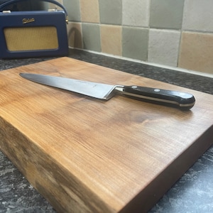 May include: A wooden chopping board with a natural wood grain finish. A silver knife with a black handle is lying on the board.