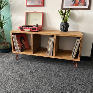 May include: A wooden record player stand with three compartments, holding vinyl records. A red record player sits on top. The stand has copper-coloured hairpin legs and is against a white wall with framed art.
