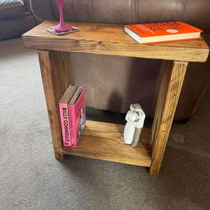 May include: A rustic wooden side table with a shelf. The table has a natural wood finish and is decorated with a glass of wine, a book, and a figurine. The table is sitting on a grey carpet.