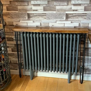 May include: A dark grey column radiator featuring a rectangular wooden shelf top supported by four black metal hairpin legs. The radiator has vertical fins and is positioned against a wall with a light brown and white brick-patterned wallpaper.