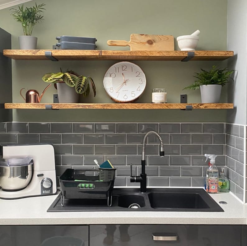May include: Two wooden shelves with black metal brackets, mounted on a gray tiled wall. The shelves are decorated with plants, a clock, and a candle. A black double sink with a chrome faucet is below the shelves.