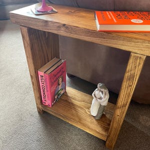 May include: A wooden side table with a shelf. The table has a natural wood finish and is decorated with a book, a small statue, and a pink glass. The book is titled "The Subtle Art of Not Giving a F*ck" by Mark Manson.