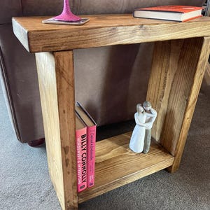 May include: A rustic wooden side table with a shelf. The table has a natural wood finish and is stained a dark brown colour. There are two books and a small figurine on the shelf. The books are titled "Billy Connolly" and "Windswept & Interesting".