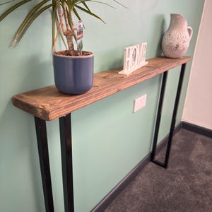 May include: A slimline wooden console table with black metal legs. A potted plant in a blue pot, a white "HOME" sign, and a speckled ceramic jug sit on the table. The table is against a pale green wall.