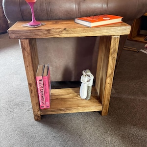 A wooden side table with a pink wine glass, a book, and a small white figurine on it, placed on a gray carpeted floor.
