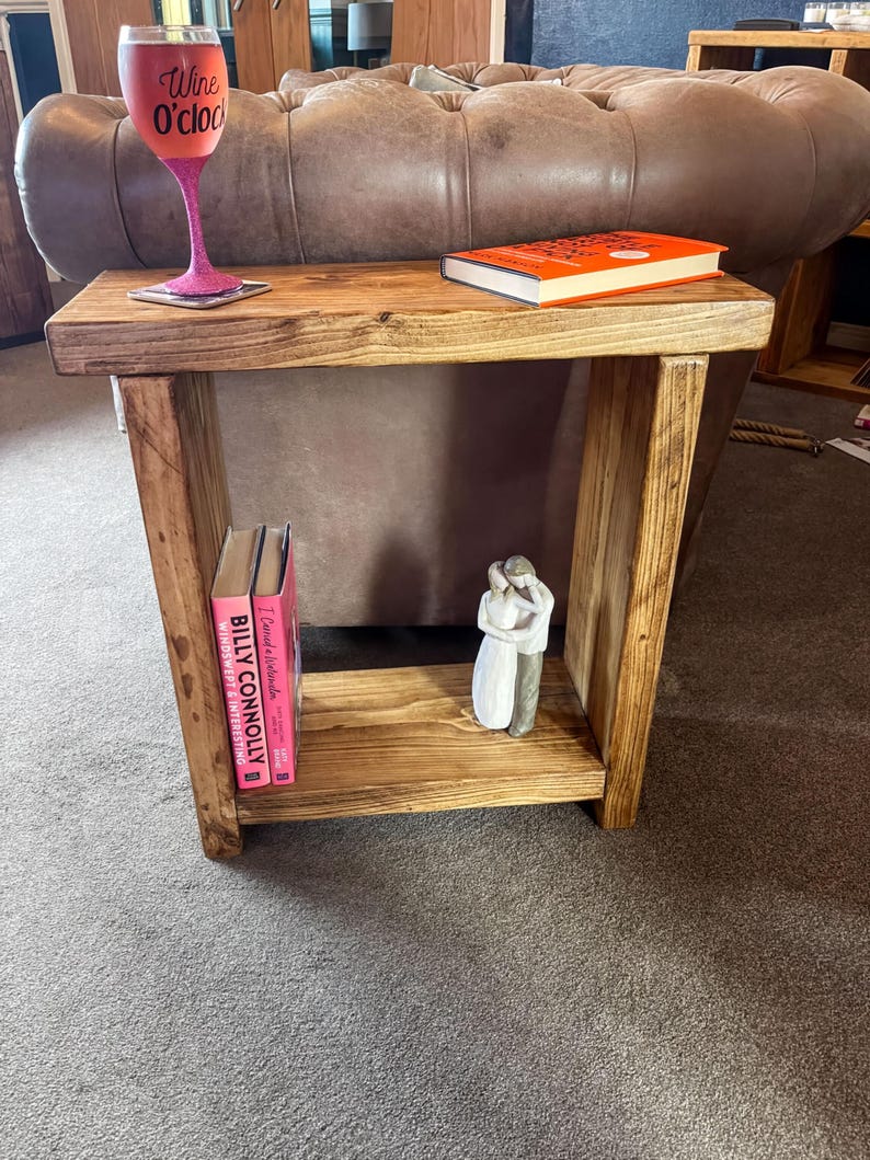 A wooden side table with a pink wine glass, a book, and a small white figurine on it, placed on a gray carpeted floor.
