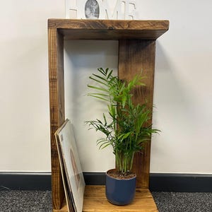 May include: A rustic wooden shelf unit with a single shelf and a small open space below. The shelf is decorated with a framed photo, a potted plant, and a stack of vinyl records. The shelf is made of reclaimed wood and has a natural, distressed finish.