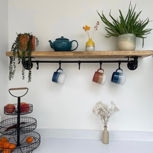 May include: A rustic wooden shelf with black metal pipe brackets and hooks. The shelf is decorated with a teal teapot, two mugs with blue and white stripes, a mug with orange and white stripes, a small potted plant, a vase with dried flowers, and a potted succulent.