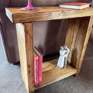 a wooden end table with a pink vase, a book, and a figurine on it, placed on a gray carpeted floor.