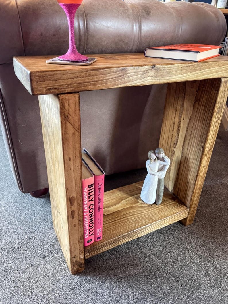 a wooden end table with a pink vase, a book, and a figurine on it, placed on a gray carpeted floor.