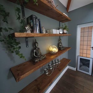 May include: Rustic wooden shelves with black metal brackets, decorated with books, statues, and candles. The shelves are mounted on a grey wall with a white trim.