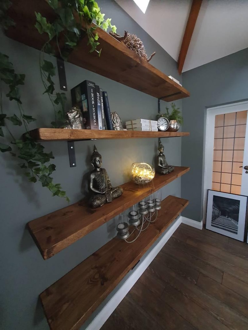 May include: Rustic wooden shelves with black metal brackets, decorated with books, statues, and candles. The shelves are mounted on a grey wall with a white trim.