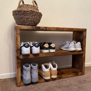May include: A wooden shoe rack with three shelves. The top shelf is empty, the middle shelf has two pairs of white sneakers with black accents and the text "Nike Air" on the side, and the bottom shelf has two pairs of brown and gray boots.