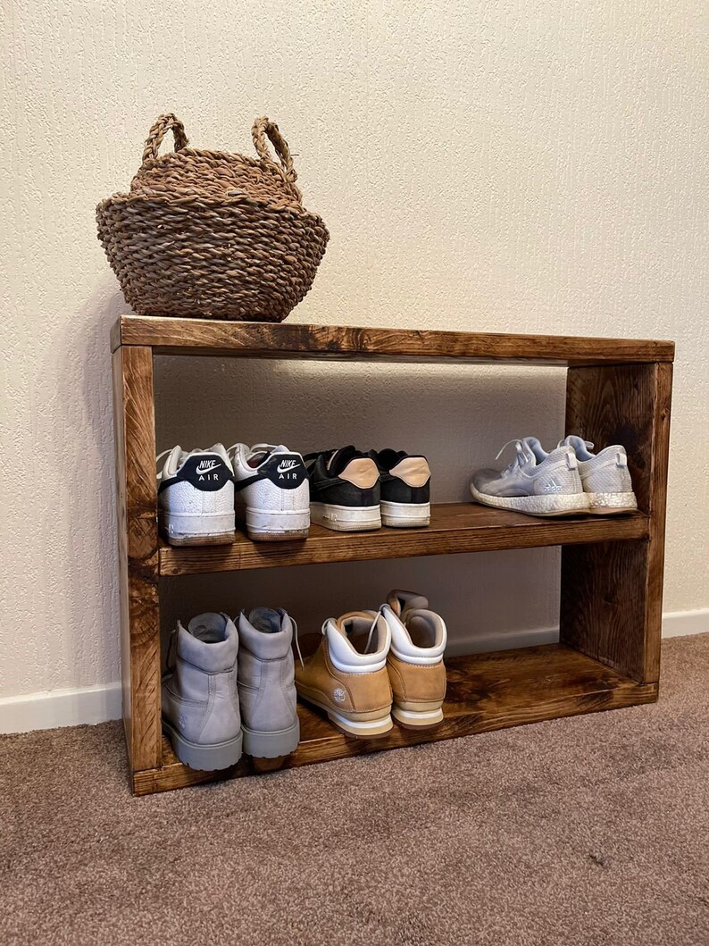 May include: A wooden shoe rack with three shelves. The top shelf is empty, the middle shelf has two pairs of white sneakers with black accents and the text "Nike Air" on the side, and the bottom shelf has two pairs of brown and gray boots.