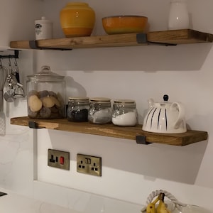 Puede incluir: Dos estantes de madera con soportes met&aacute;licos, montados en una pared blanca. El estante superior tiene un jarr&oacute;n de cer&aacute;mica amarillo y un cuenco naranja. El estante inferior tiene un tarro de vidrio lleno de patatas, tres tarros de vidrio con tapas y una tetera blanca con rayas azules.