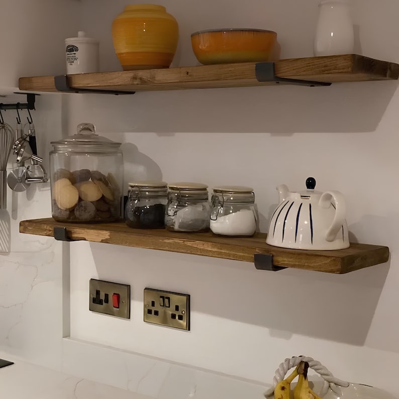 Puede incluir: Dos estantes de madera con soportes met&aacute;licos, montados en una pared blanca. El estante superior tiene un jarr&oacute;n de cer&aacute;mica amarillo y un cuenco naranja. El estante inferior tiene un tarro de vidrio lleno de patatas, tres tarros de vidrio con tapas y una tetera blanca con rayas azules.