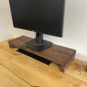 May include: A dark stained wooden monitor stand with a black computer monitor on top. A black keyboard is placed on the stand, and a black computer mouse is on the wooden desk. The monitor stand is approximately 76 cm long.