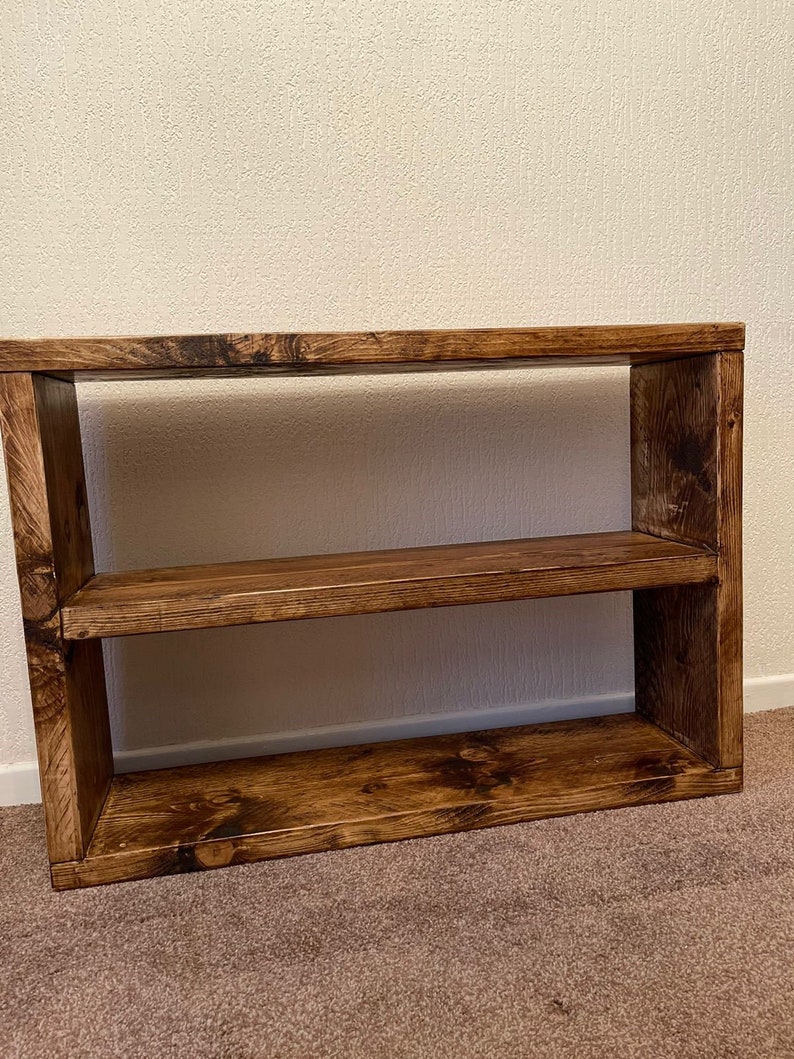May include: A rustic wooden shelf unit with two shelves. The shelf is made of dark brown wood with a natural wood grain finish.