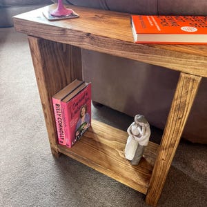 A wooden side table with a pink book and a small figurine on it, placed on a carpeted floor next to a couch.