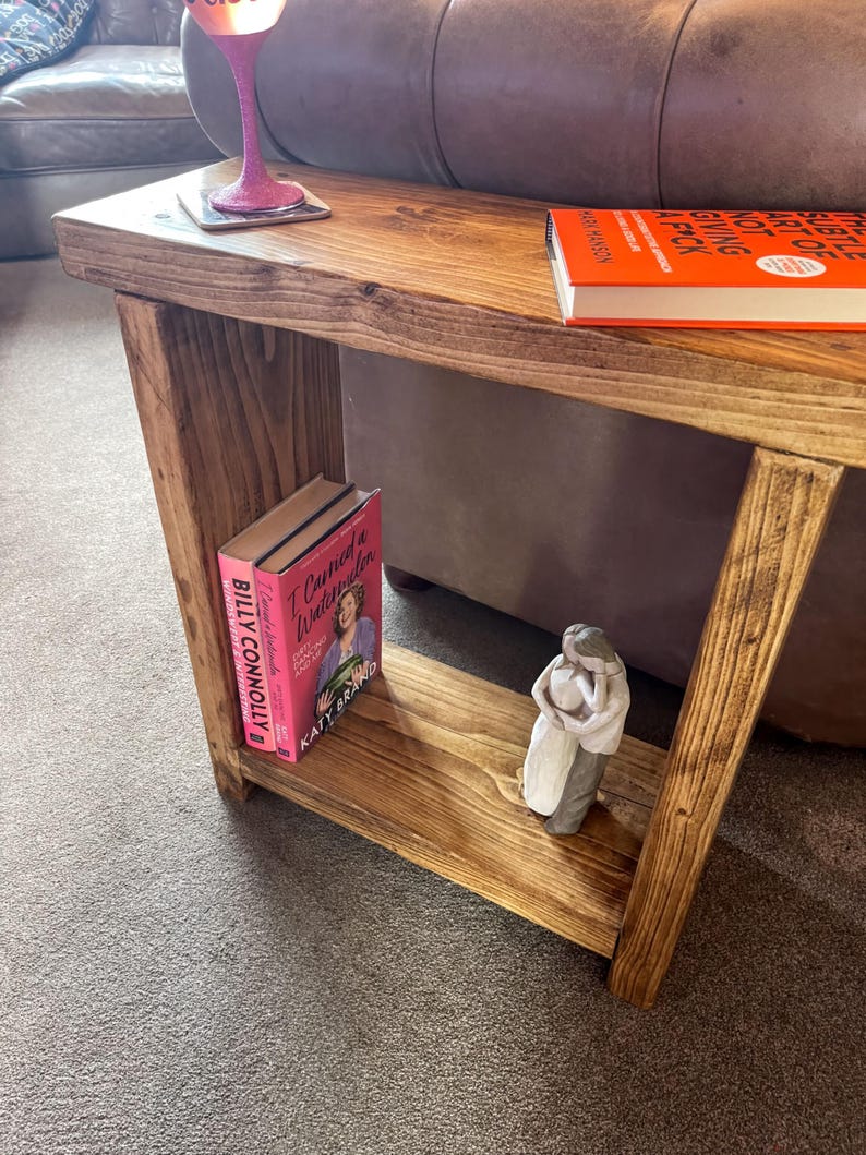 A wooden side table with a pink book and a small figurine on it, placed on a carpeted floor next to a couch.