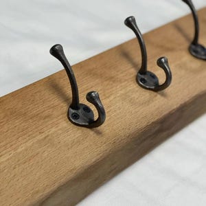 May include: A rectangular light brown wooden board featuring three dark grey metal hooks. Each hook is secured to the wood with two small black screws. The hooks have a curved, double-pronged design with rounded tips for hanging items.