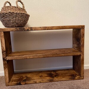 May include: A wooden shelf unit with two shelves. The shelf is stained a dark brown color and has a rustic look. A woven basket sits on top of the shelf.