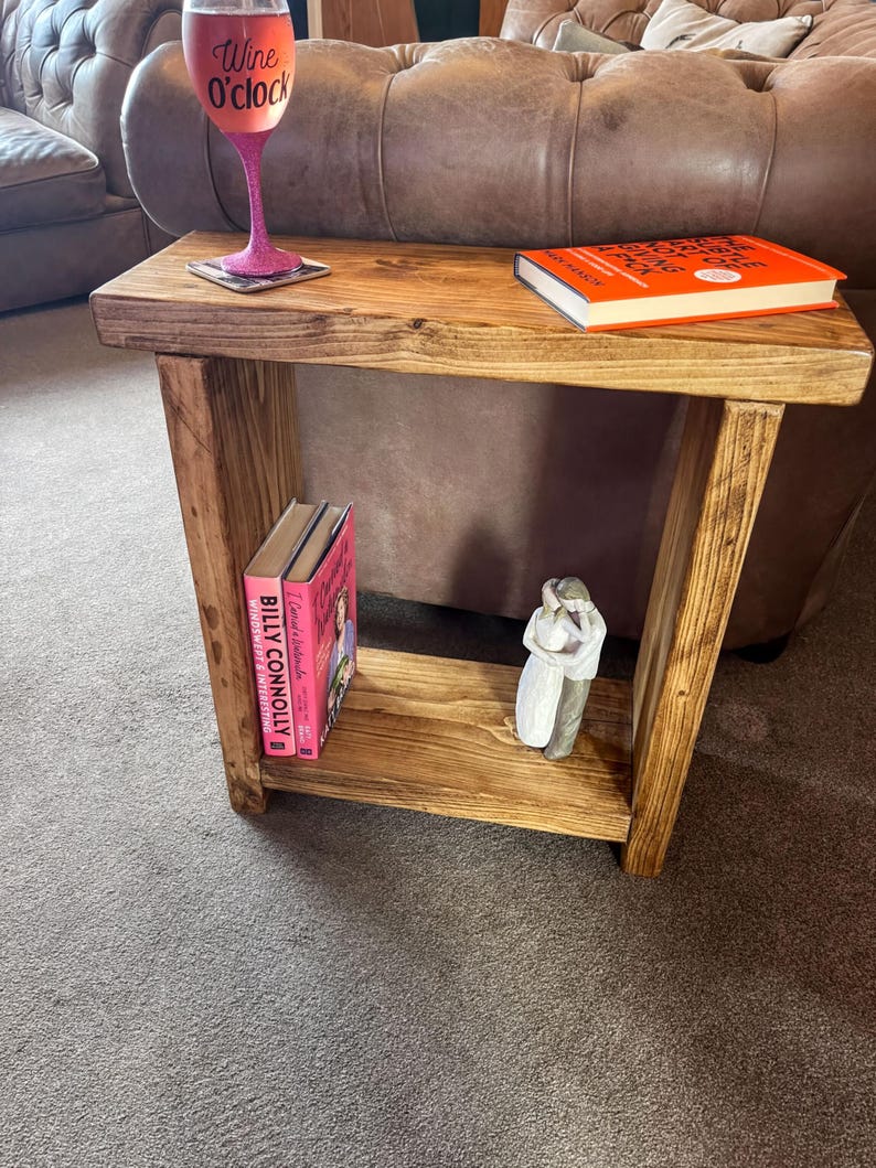 A wooden side table with a pink wine glass and a book on it, placed in a living room setting with a brown leather couch in the background.
