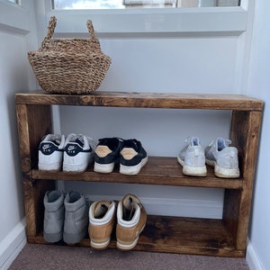 May include: A wooden shoe rack with two shelves. The rack is stained a dark brown color and has a rustic look. There are six pairs of shoes on the shelves, including sneakers, boots, and sandals.
