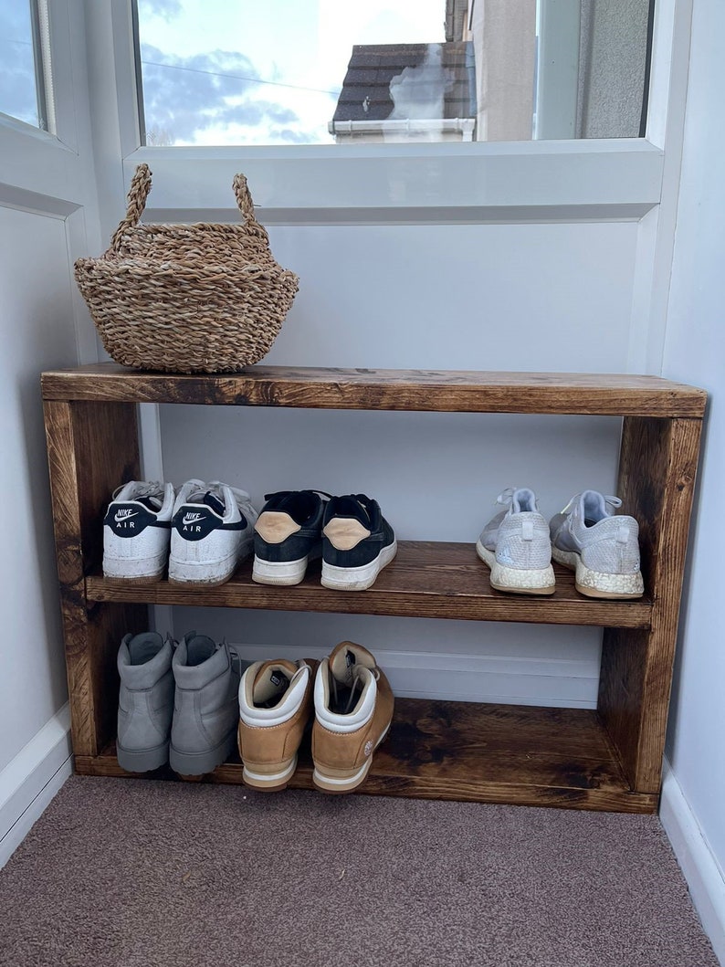 May include: A wooden shoe rack with two shelves. The rack is stained a dark brown color and has a rustic look. There are six pairs of shoes on the shelves, including sneakers, boots, and sandals.