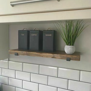 May include: A wooden shelf with black metal brackets, holding three black canisters labeled "Tea", "Coffee", and "Sugar". The shelf is mounted on a white tiled wall.