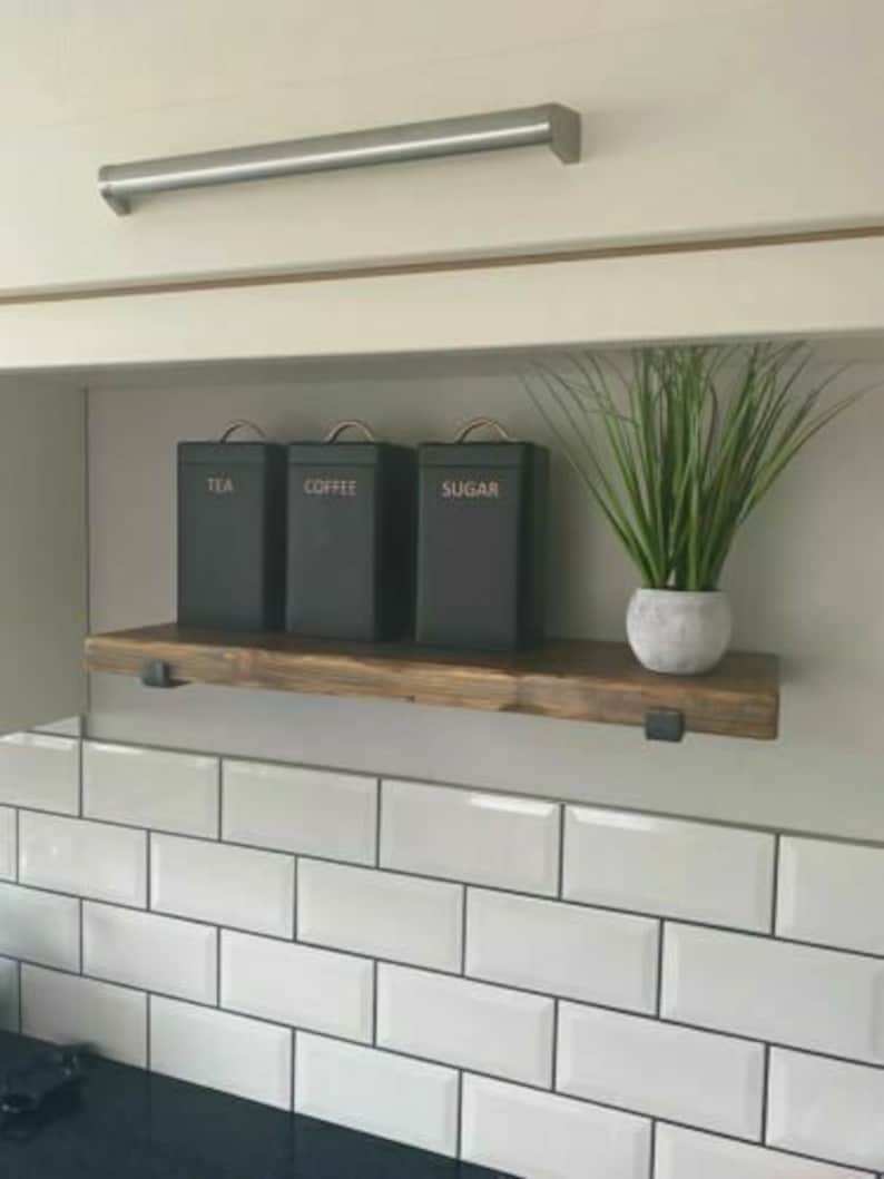 May include: A wooden shelf with black metal brackets, holding three black canisters labeled "Tea", "Coffee", and "Sugar". The shelf is mounted on a white tiled wall.