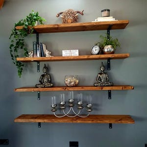 May include: Four wooden shelves with black metal brackets mounted on a grey wall. The shelves are decorated with plants, books, candles, a small animal head sculpture, and two small statues. The text on the books reads "Take time to do what makes your soul happy."