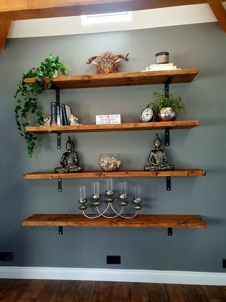 May include: Four wooden shelves with black metal brackets mounted on a grey wall. The shelves are decorated with plants, books, candles, a small animal head sculpture, and two small statues. The text on the books reads "Take time to do what makes your soul happy."