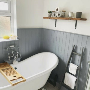 May include: A white bathtub with a wooden bath tray, a chrome faucet, and a gray tiled wall. A wooden shelf with black metal brackets is mounted on the wall above the tub. The shelf holds a small potted plant, a white ceramic candle, and a wire basket with toilet paper rolls. A gray ladder with two white towels is leaning against the wall to the right of the tub.
