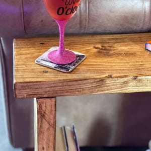 A pink glitter wine glass with the text "Wine O'clock" sits on a wooden side table, accompanied by a coaster and a stack of books.
