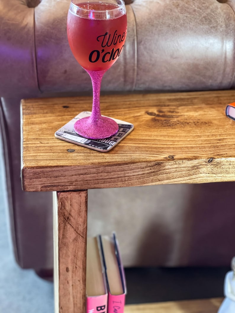 A pink glitter wine glass with the text "Wine O'clock" sits on a wooden side table, accompanied by a coaster and a stack of books.