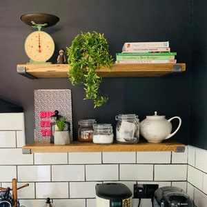 Puede incluir: Dos estantes de madera con soportes de metal negro, montados en una pared de azulejos blancos. El estante superior tiene una b&aacute;scula verde vintage, una figurita de madera y una planta en maceta. El estante inferior tiene un cuaderno con estampado en blanco y negro, una peque&ntilde;a planta en maceta, tres frascos de vidrio y una tetera blanca con dise&ntilde;o floral.