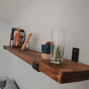 May include: A rustic wooden shelf with black metal brackets. The shelf is decorated with a glass of water, a potted plant, and a few books.
