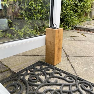 May include: A wooden door stop with a metal handle, positioned to hold a door open. The door stop is made of light brown wood and sits on a stone patio. A black decorative doormat is in the foreground. The door stop is approximately 20 cm tall.