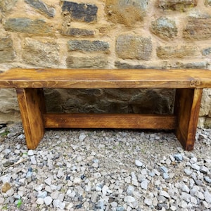 May include: A rustic wooden bench with a natural wood finish. The bench has a simple design with a single plank seat and two legs on each side. The bench is sitting on a gravel surface.