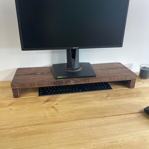 May include: A dark brown wooden monitor stand supporting a black computer monitor. A black keyboard sits on the stand. The stand is on a light brown wooden desk.