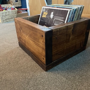 May include: A wooden record storage box with a dark brown stain. The box is filled with vinyl records, including one with the text "Billy Joel" on the cover.