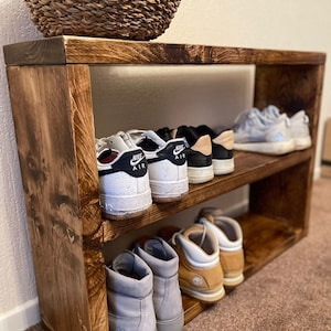 May include: A two-tier wooden shoe rack with a natural wood finish. The top shelf has three pairs of sneakers, including two pairs of white Nike Air Force 1s. The bottom shelf has three pairs of brown and gray boots.