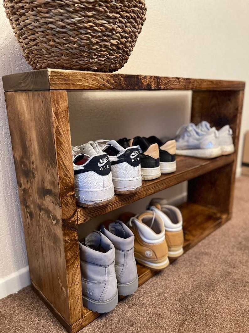 May include: A two-tier wooden shoe rack with a natural wood finish. The top shelf has three pairs of sneakers, including two pairs of white Nike Air Force 1s. The bottom shelf has three pairs of brown and gray boots.